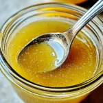 Homemade lemon vinaigrette in a glass jar with fresh lemons, garlic, and herbs on a wooden table.